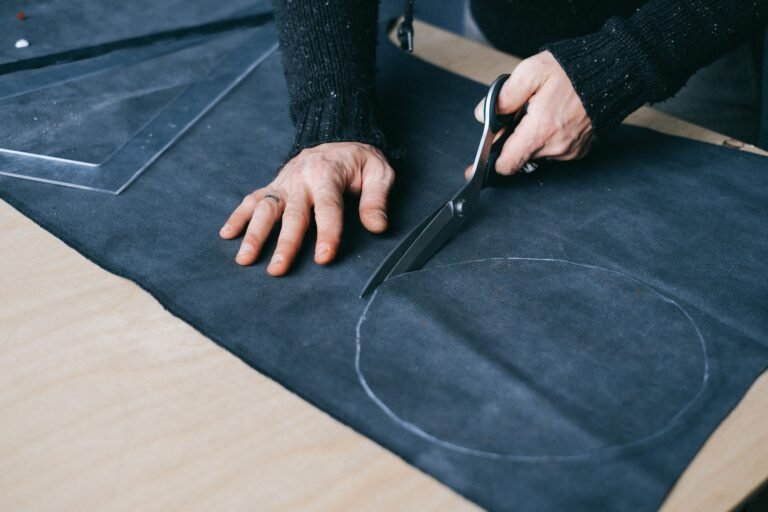 High angle of crop anonymous male tailor with scissors cutting round piece of cloth while working in atelier